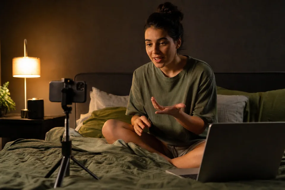 Adult creator filming a clip with her phone on a tripod in a dim bedroom